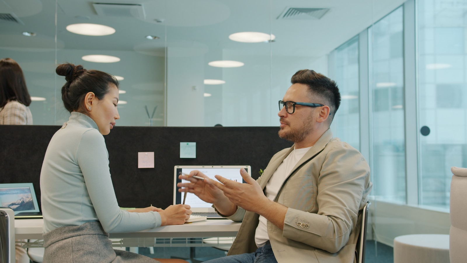 An Asian woman taking notes during a serious 1:1 conversation with a colleague at a desk in a modern office, the shape of the conversation this article is about.