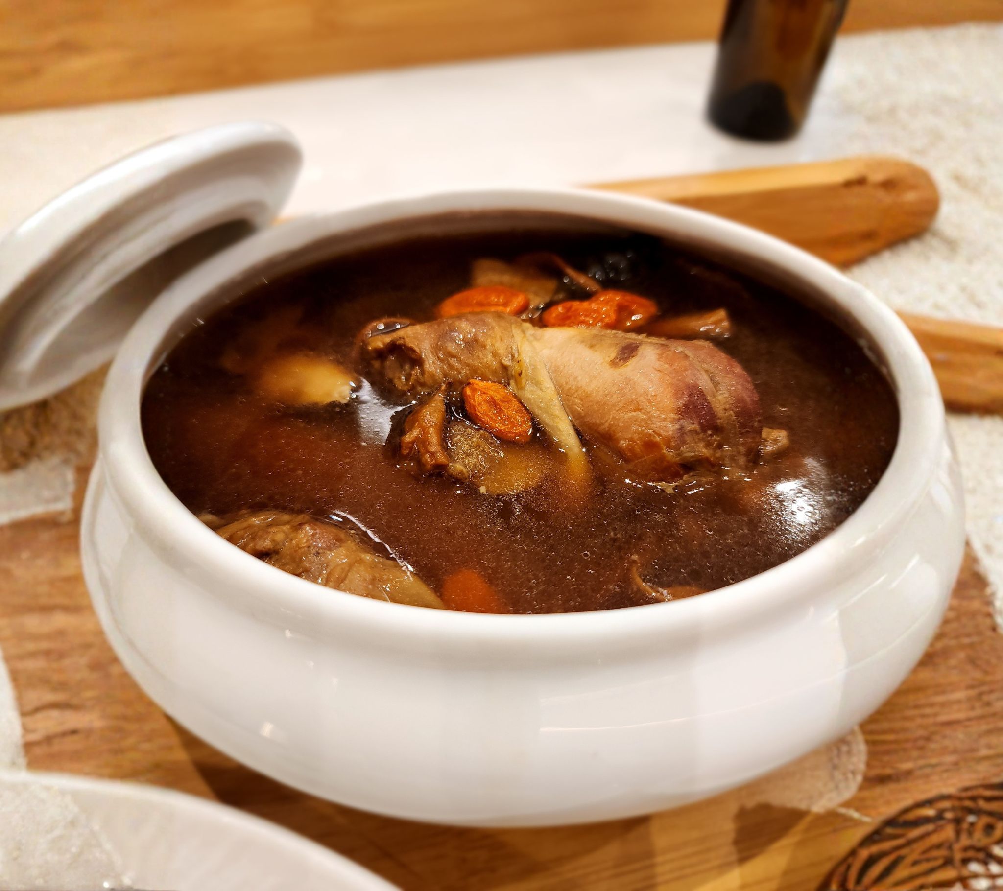 A white tureen of Chinese herbal chicken soup with goji berries, ginger and red dates floating in a clear dark broth