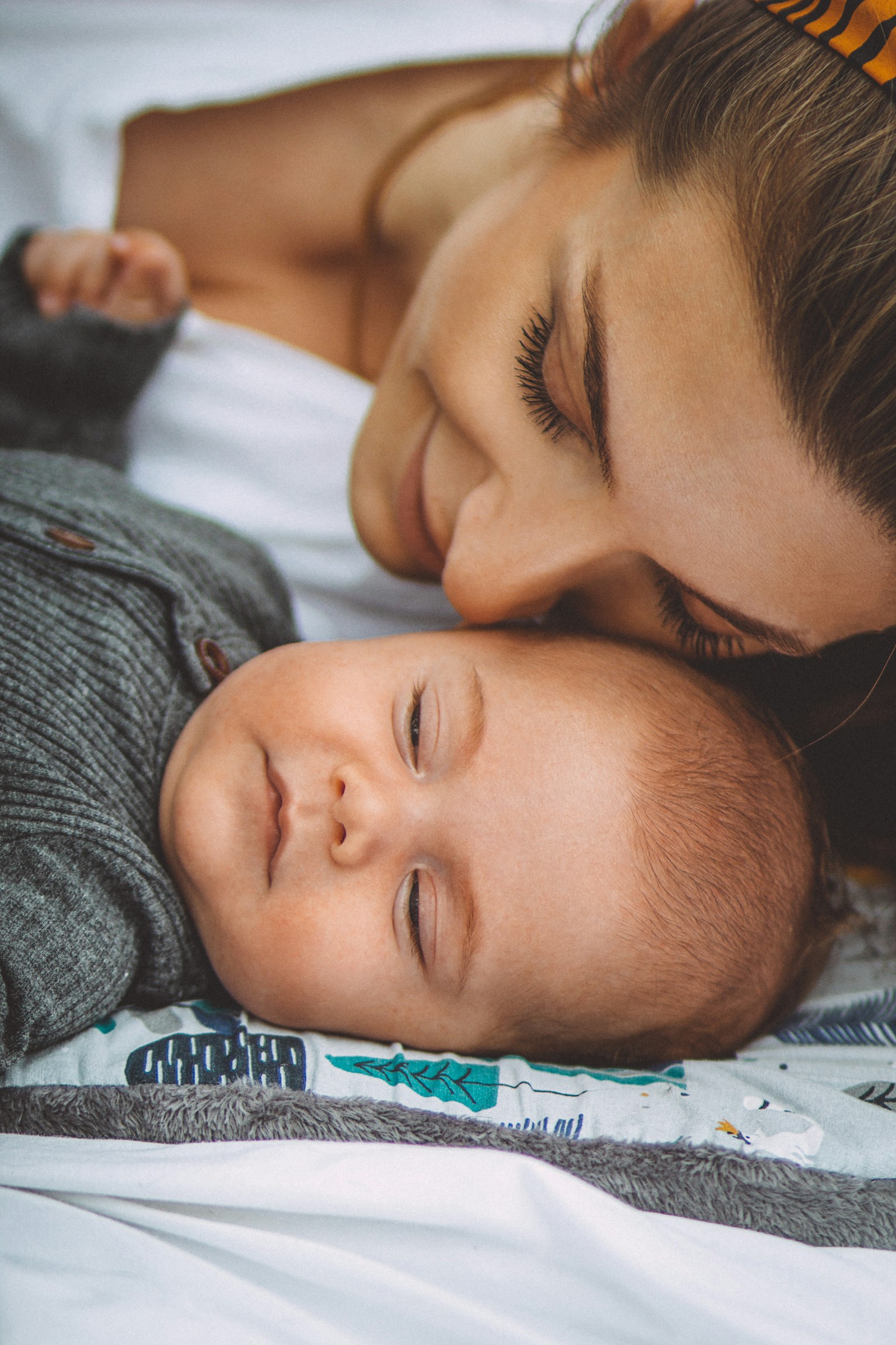 A mother resting on her side in bed next to her sleeping baby, the quiet posture of a night feed and a rest that ran into a doze.