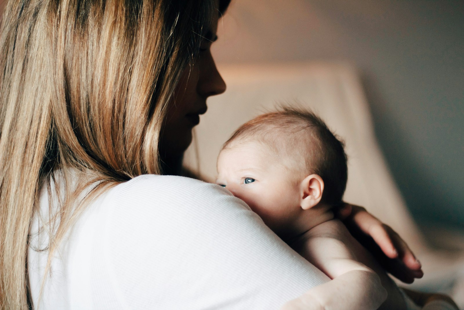 A young mother in a white shirt holds her baby close as they move through a quiet space, the kind of constant carrying that doubles for the invisible mental work she is also doing