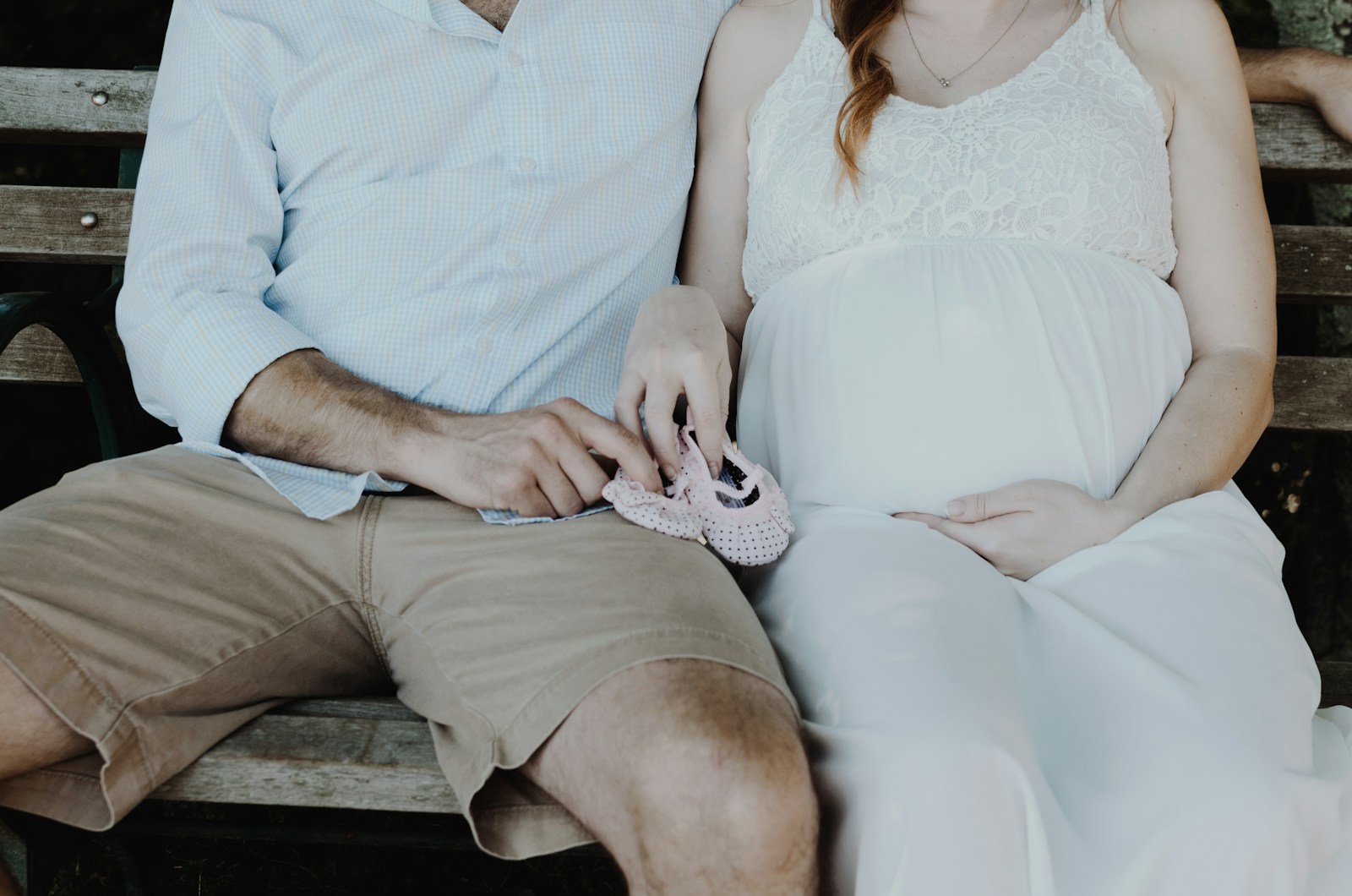 A pregnant woman and her partner sit close together on a wooden bench, his hand resting on her bump as she holds a tiny pair of pink baby shoes, the two of them thinking through what comes next